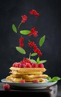 Stack of belgian waffles in a plate with raspberries and red currants on a black background, green mint leaves levitate photo