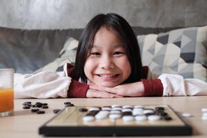 Portrait Close-up shot, one Asian girl fun playing Go, checkers board game on table in home living room, cheerful smile and looking at camera, happy weekend relaxing hobby of clever cute child winner. photo