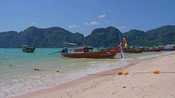 Traditional long tail boats with colorful ribbons dock along a sandy beach in Phi Phi Islands, framed by turquoise waters and limestone cliffs. video