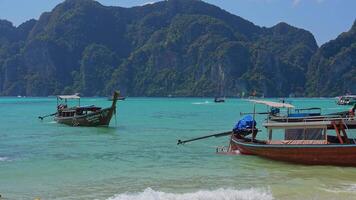 Traditional longtail boats float near limestone cliffs and turquoise waters, with lush greenery and a clear blue sky in Phi Phi Islands, Thailand. video