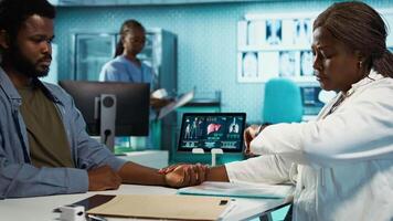 Physician measuring the heart rate of a young man by using her smartwatch, employing the digital fitness tracker accessory on her gadget. Patient and doctor interaction with tools. Camera B. video