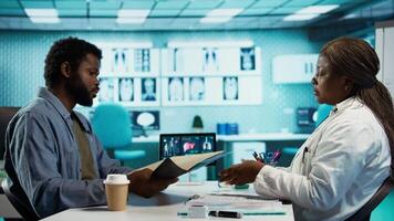 Black male patient bringing x ray test results to medic for scan interpretation, reviewing the reports for diagnostics. Medic expert evaluating the radiography film in cabinet. Camera B. video
