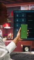 Vertical Woman seated at desk using AI LLM chatbot software and green screen phone in modern urban home office. Person uses large language model program on computer and mockup smartphone in apartment, camera A video
