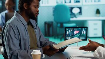 Black male patient bringing x ray test results to medic for scan interpretation, reviewing the reports for diagnostics. Medic expert evaluating the radiography film in cabinet. Camera A. video