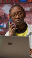 Vertical Woman in home office sitting at desk in front of laptop, answering phone call. Remote employee using notebook to do job online, receiving telephone call on smartphone, camera B video