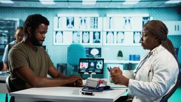 Black medic measuring the blood pressure and pulse of a male patient, doing routine checkup with a blood pressure monitor. Physician using advanced tools for cardiology readings. Camera B. video