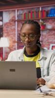 Vertical Person enjoying leisure time at home, eating snacks, texting friends on laptop. African american woman enjoying croissant, talking with mates on social network online website, using notebook, camera B video