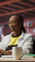 Vertical Remote employee drinking coffee at home while looking at social media posts on notebook. African american woman enjoying hot beverage and reading ebooks on laptop, camera A video