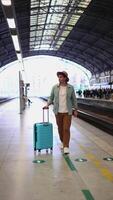 verticaal nerveus reiziger met hoed en blauw koffer wandelen Aan platform aan het wachten voor vertraagd trein video