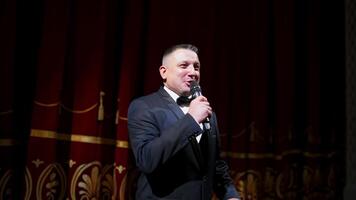 Kyiv, Ukraine, 2 May 2024 Male announcer with microphone on the stage. Actor in black suit on red scene curtains background. video