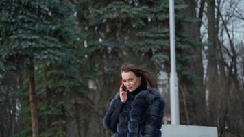 luxe fille avec une téléphone dans le parc. portrait de attrayant Dame dans fourrure manteau en marchant dans le parc et parlant le mobile téléphone. lent mouvement. video