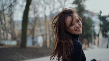 Happy girl turning around in the park. Attractive young lady with long hair in black sweater looking on camera and smiling in the street of a city. Slow motion. video