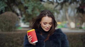 Portrait of a smiling woman. Attractive brunette girl in fur coat stands with plastic cup of coffee in the park and looking on camera. Slow motion. video