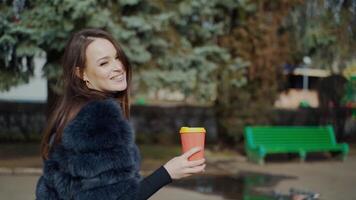 Attractive girl posing on camera. Pretty young woman in black fur coat standing in the park with coffee and smiling on camera. video