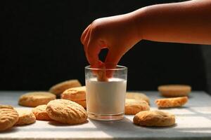 Child dipping tasty cookie in glass of milk on black background photo