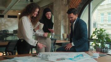 Multiethnic designers engineers architects business coworkers Caucasian Muslim women Indian man talk discuss plan architecture blueprints on table friendly conversation in office drink coffee teamwork video