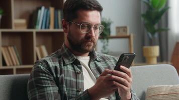 Cheerful calm serious relaxed man at home guy male gamer user using mobile phone smartphone device gadget addict chatting social media shopping online app net at living room at home couch browsing video
