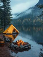 A serene lakeside camping scene with a yellow tent, campfire, and misty mountains. photo