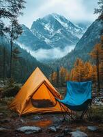 A serene camping scene with an orange tent and blue chair surrounded by mountains and trees. photo