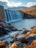 A serene waterfall cascades over a stone dam, surrounded by autumn foliage and rocky terrain. photo