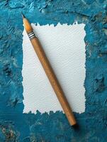 A pencil rests on a textured blue background next to a blank sheet of paper. photo