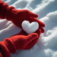 A pair of hands in red knitted gloves holding a heart-shaped snowball. The background is a snowy surface with a soft texture. photo