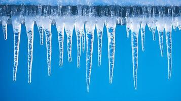 Long icicles hang from roof edge against crystal clear blue sky, displaying frozen water droplets and transparent ice formation with dramatic natural lighting in winter scene. icicle photo