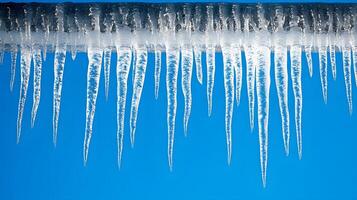 Long icicles hang from roof edge against crystal clear blue sky, displaying frozen water droplets and transparent ice formation with dramatic natural lighting in winter scene. dramatic photo