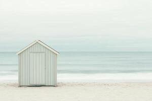 A small wooden beach hut in neutral tones stands on a smooth sandy beach, framed by soft waves and a calming, cloudy sky. This tranquil setting invites relaxation and reflection photo