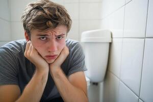 A young man sits on the bathroom floor looking worried and uncomfortable near a toilet during a distressing moment .Constipation, digestive problems concept. photo