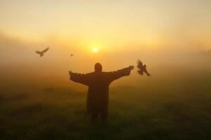 A person stands with arms outstretched at sunrise, surrounded by birds in a foggy field during early morning hours photo