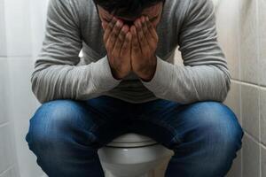 A man sitting on the toilet with his head in his hands, expressing distress or anxiety in a small bathroom setting during daytime .Constipation, digestive problems concept. photo