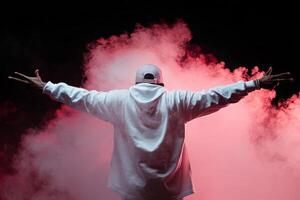 A confident individual in a white hoodie poses with arms outstretched amidst colorful smoke at a nighttime event photo
