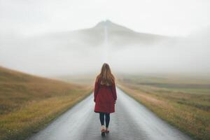 A woman in a red coat walks down a misty road surrounded by green hills and a distant waterfall on a cloudy day photo