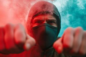 A masked individual confronts the camera surrounded by colorful smoke at an outdoor gathering during the evening photo