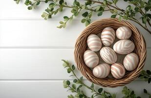 Decorative eggs in a woven basket surrounded by greenery on a white wooden surface photo