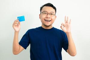 Delighted asian man hold credit card with Ok hand sign pose. Banking, Financial isolated background, blue color concept. photo