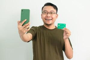 Delighted asian man wear green tshirt hold credit card and phone. Banking, Financial and technology isolated background photo