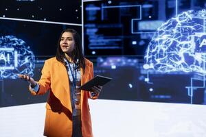 Company CEO talking at AI expo using mic and tablet, sharing industry expertise to audience. Computer scientist at tech event holding device, presenting latest AI products and services photo
