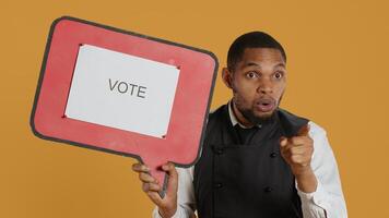 Catering employee showing a message to vote on red speech bubble, presenting the cardboard icon to do a commercial for politics. Waiter in uniform works on customer service. Camera A. photo
