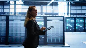 Engineer in server hub checking recovery plan on tablet, monitoring rackmounts energy consumption. Woman in data center making sure rigs are functioning optimally, doing routine disk checks, camera B photo