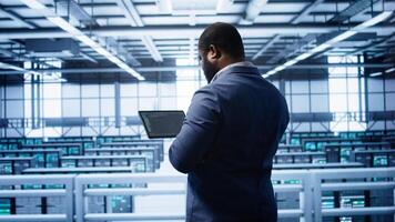Computer scientist in data center running error checking utilities on tablet to fix bugs. IT expert in server hub facility looking for network issues affecting supercomputers performance, camera B photo