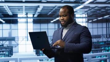 Programmer in data center typing on laptop, running error checking utilities. IT professional in server room looking for network issues affecting rigs performance using digital device, camera A photo