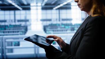 System administrator monitoring server room, doing checkup, inputting data on tablet of needed fixes. IT support employee working in data center facility housing storage rackmounts, camera B close up photo