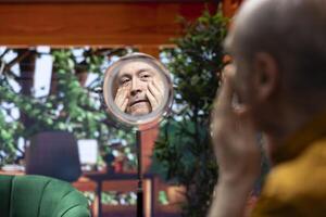 Pensioner using serum and cream to preserve his mature skin with wrinkles, looking in the mirror at his aging face. Doing a pampering daily routine for personal care, grooming products. photo