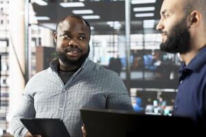 Multiracial businesspeople in startup company looking at business documents on tablet and laptop. Coworkers in office checking crosschecking figures on device and notebook, talking about data photo