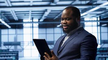 Programmer in server room location using tablet to patch network issues affecting transfer rates. African american man in data center fixing errors affecting mainframes, camera A photo