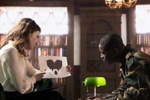 Female therapist presents a Rorschach inkblot test to a black soldier in armed forces uniform during a counseling session. Diverse people at therapy in a warm office with bookshelves. photo