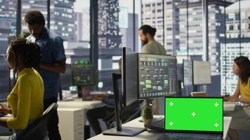 Green screen laptop on desk in workspace with admins in background doing software quality assurance. Chroma key notebook in office with woman at work inspecting coding on device, camera B photo