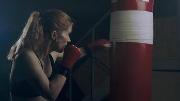 Female fighter in darkened room at home in garage practicing fast strong punches on punching bag, boxing gym training in slow motion, 4k video